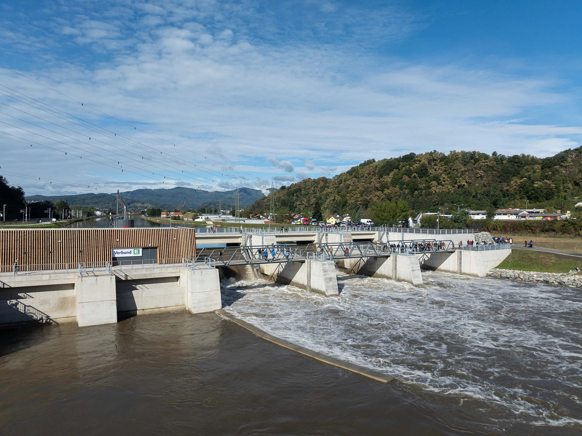 Wir blicken auf das Laufkraftwerk Gratkorn bei Sonnenschein. Auf Fußgängerbrücke sind zahlreiche Menschen zu sehen.