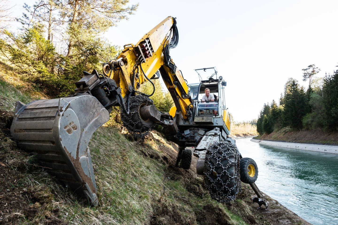 Ein Bagger arbeitet an der Baustelle zur Erneuerung des VERBUND Kraftwerks Jettenbach-Töging. Der gelbe Bagger steht auf einer Böschung neben einem Kanal und ist in Aktion, während ein Arbeiter in der Fahrerkabine sitzt.