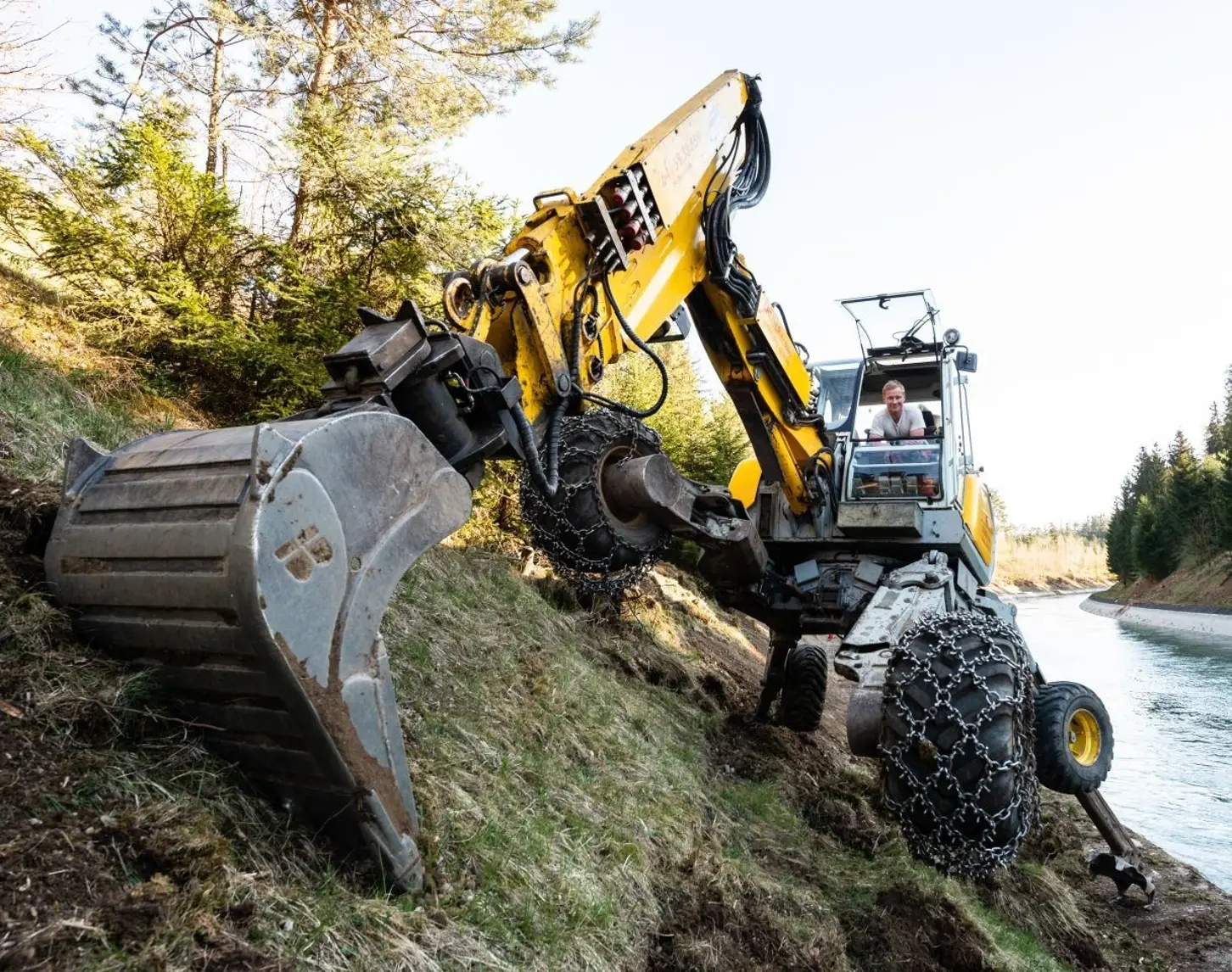 Ein Bagger arbeitet an der Baustelle zur Erneuerung des VERBUND Kraftwerks Jettenbach-Töging. Der gelbe Bagger steht auf einer Böschung neben einem Kanal und ist in Aktion, während ein Arbeiter in der Fahrerkabine sitzt.