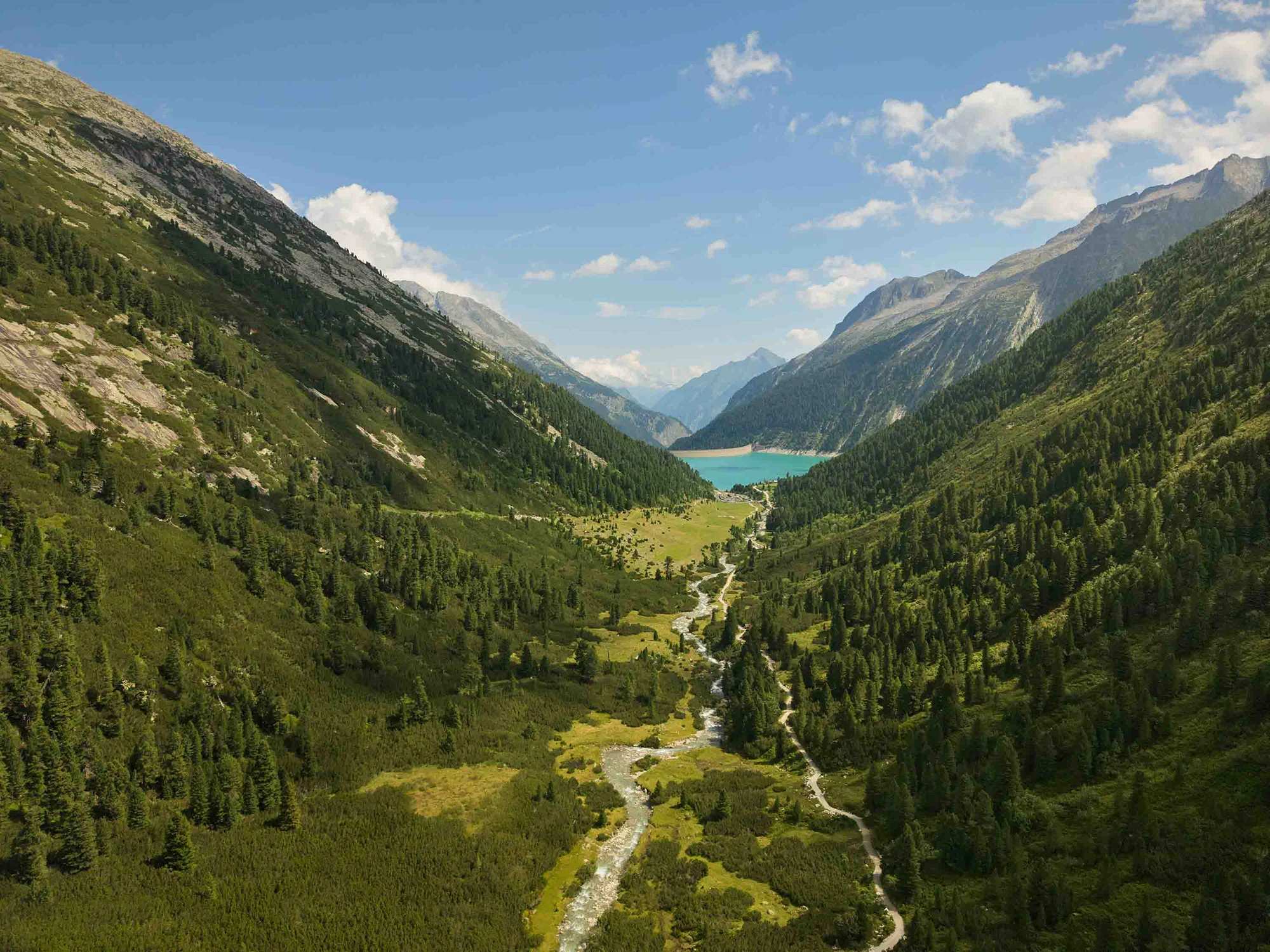 Blick über ein alpines Tal mit Fluss, Wanderweg und bewaldeten Berghängen; im Hintergrund liegt ein türkisblauer Stausee.