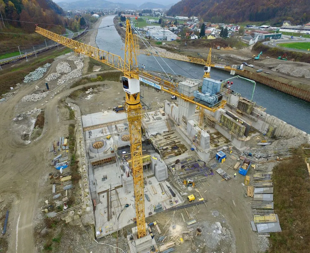Luftbild der Kraftwerksbaustelle am Ufer der Mur. Auf dem Bild ist eine Mischung aus Bagger, Baumaschinen und teils fertiggestellten Betonstrukturen zu sehen. Es gibt deutlich erkennbare, umgeschichtete Erdmassen und frisch angelegte Wasserwege, die durch das Baufeld führen. Trotz des industriellen Charakters der Szene sind Sicherheitszäune und Baufahrzeuge ordentlich angeordnet, was ein organisiertes Bauprojekt zeigt.