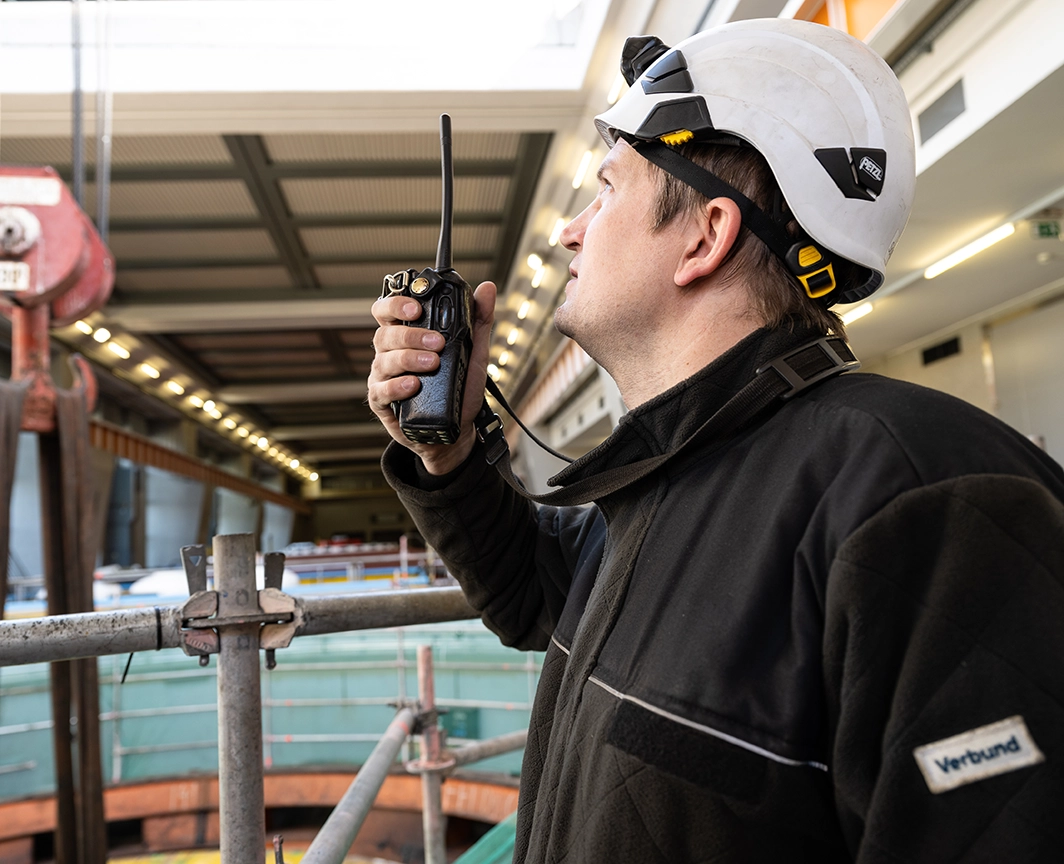 Ein VERBUND-Mitarbeiter in Schnutzkleidung und Helm blick nach oben. In der Hand hält er ein Wakie Talkie. Im Hintergrund sieht man das Innere eines Wasserkraftwerks.