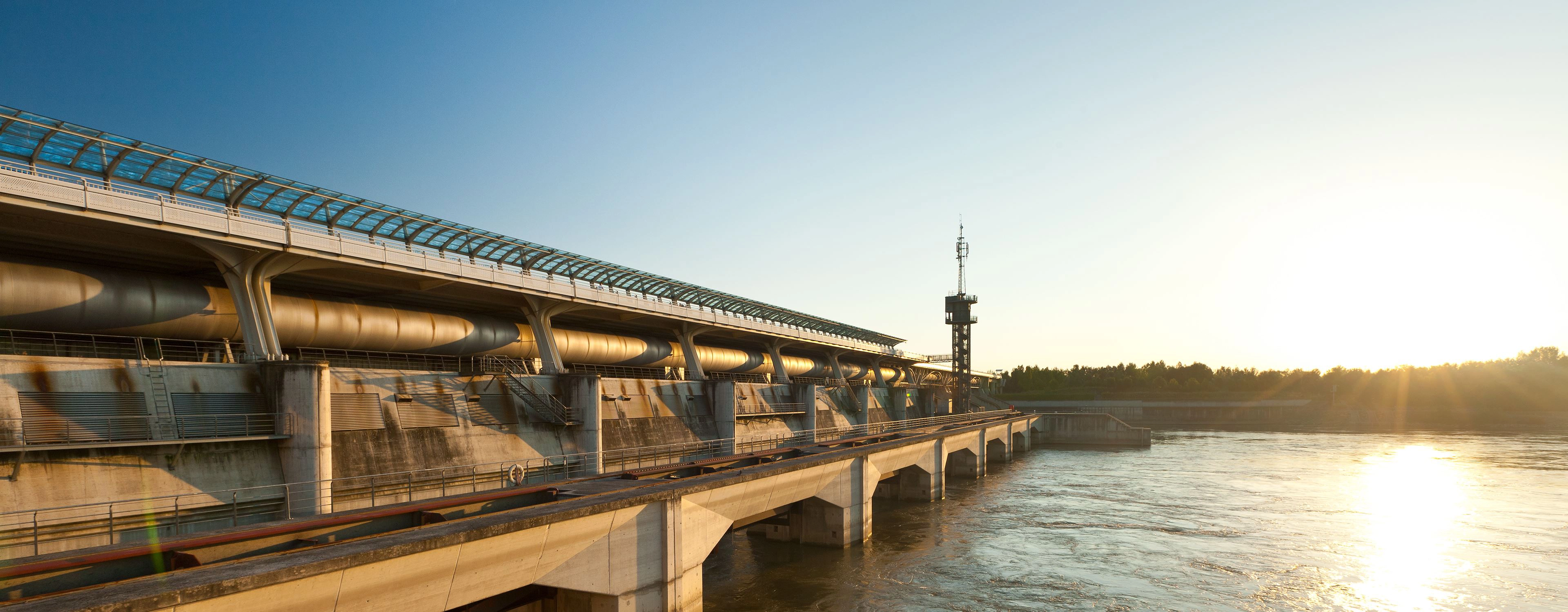 Wir blicken auf das Donaukraft Freudenau bei Wien. Die Sonne geht gerade unter und spiegelt sich im Wasser.