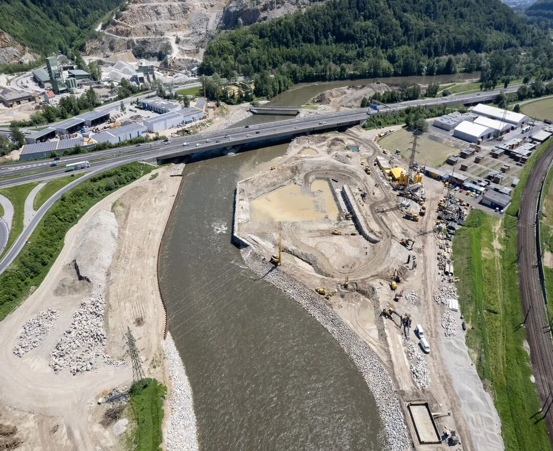 Luftbild der Baustelle in einem frühen Stadium des Projekts am Ufer der Mur. Schwere Baumaschinen, darunter Bagger und Lastwagen, sind auf einer großflächigen Baustelle zu sehen, die von Erdhaufen und ausgehobenen Gruben geprägt ist. Ein Teil des Geländes wurde für Infrastrukturarbeiten nivelliert, während in einem anderen Bereich eindeutig Erdaushub stattfindet. In der Nähe fließt der Fluss, und im Hintergrund sind eine Brücke, Bahngleise und diverse Gebäude zu erkennen, was auf eine städtische Nähe hindeutet.