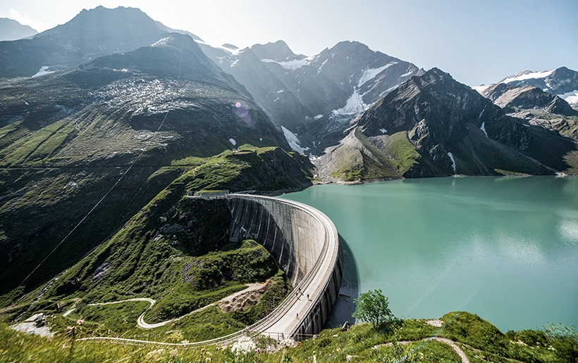 Wir blicken von oben auf das Pumpspeicherkraftwerk Kaprun. Die Wiesen sind saftig grün und die Sonne blinzelt über die Berge.