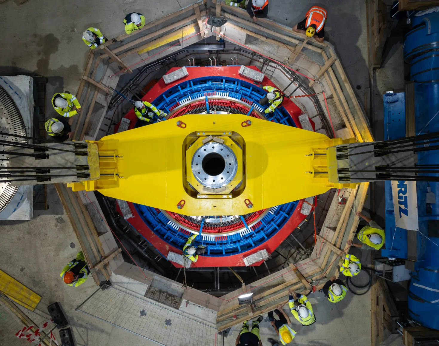 Mehrere Arbeiter in Schutzkleidung montieren in einer Halle ein grosses gelbes Bauteil über einer runden rot-blauen Maschine.