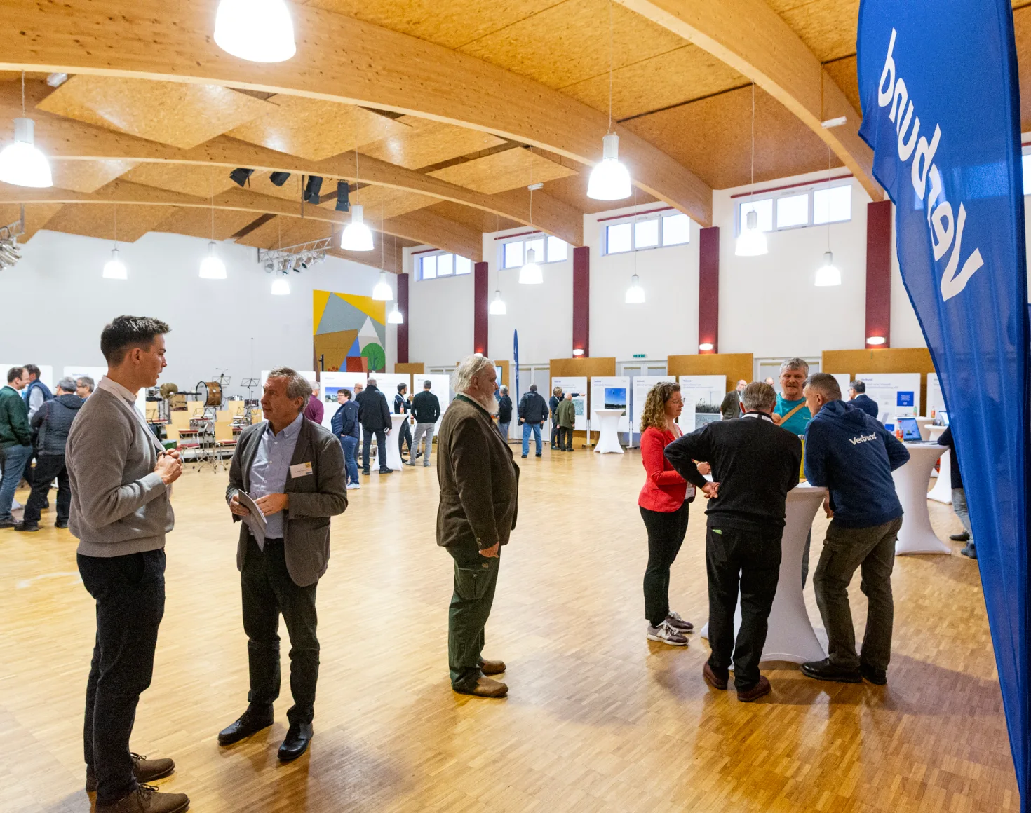 Menschen stehen in einem hellen Saal mit Holzboden und Deckenkonstruktion aus Holz. Sie führen Gespräche an Stehtischen und informieren sich an Präsentationstafeln. Ein blauer Verbund-Banner ist im Vordergrund sichtbar. Die Szene zeigt den Bürgerdialog zum Windkraftprojekt in Burgschleinitz Kühnring.
