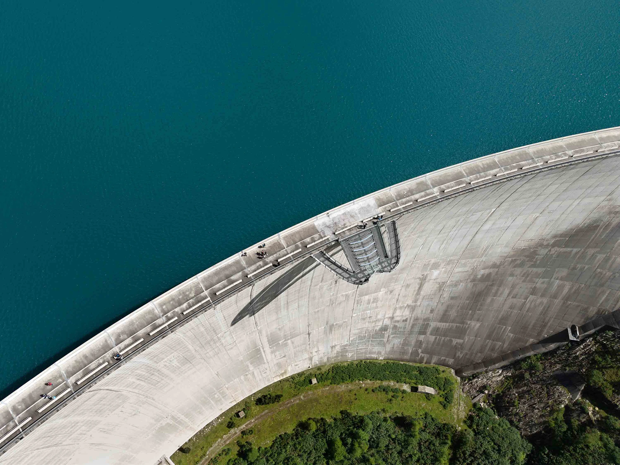 Luftaufnahme einer geschwungenen Betonstaumauer zwischen türkisblauem Stausee und bewachsenem Gelände; mehrere Menschen sind auf dem Damm zu sehen.