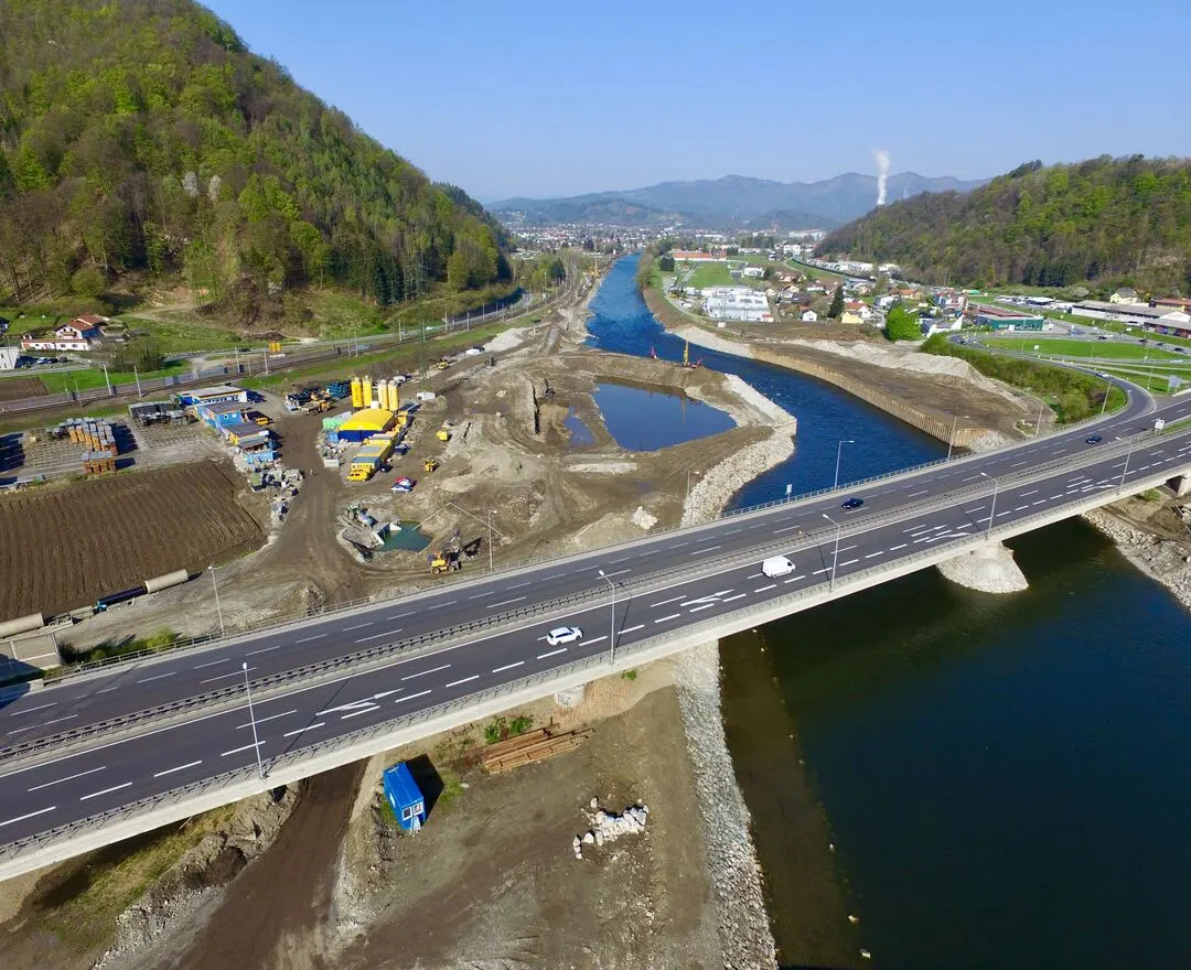 Luftbild der Baustelle im Frühstadium neben der Mur. Die Aufnahme die Straßenbrücke im Vordergrund, die über den Fluss führt. Bauarbeiten finden entlang des Flusses statt, wobei Erdbewegungsmaschinen und -fahrzeuge sichtbar sind. Im Hintergrund windet sich der Fluss durch die hügelige Landschaft, und fernere Gebäude sind am Horizont zu erkennen.