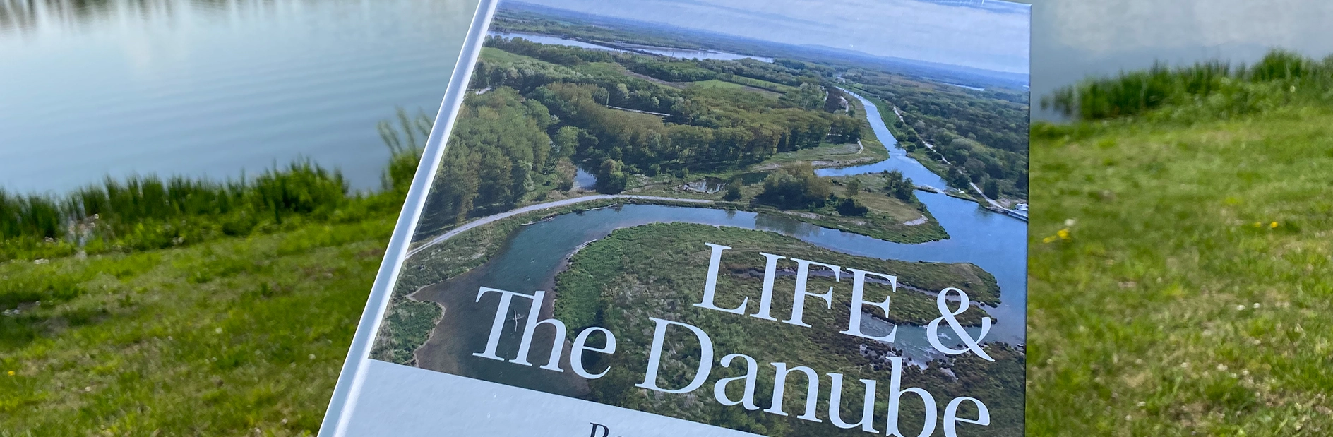 Das Bild zeigt das Buch "LIFE & The Danube", das sich mit EU-geförderten LIFE-Projekten von VERBUND beschäftigt. Auf dem Buchcover ist eine Luftaufnahme einer Flusslandschaft zu sehen. Das Buch liegt im Gras neben einem Gewässer