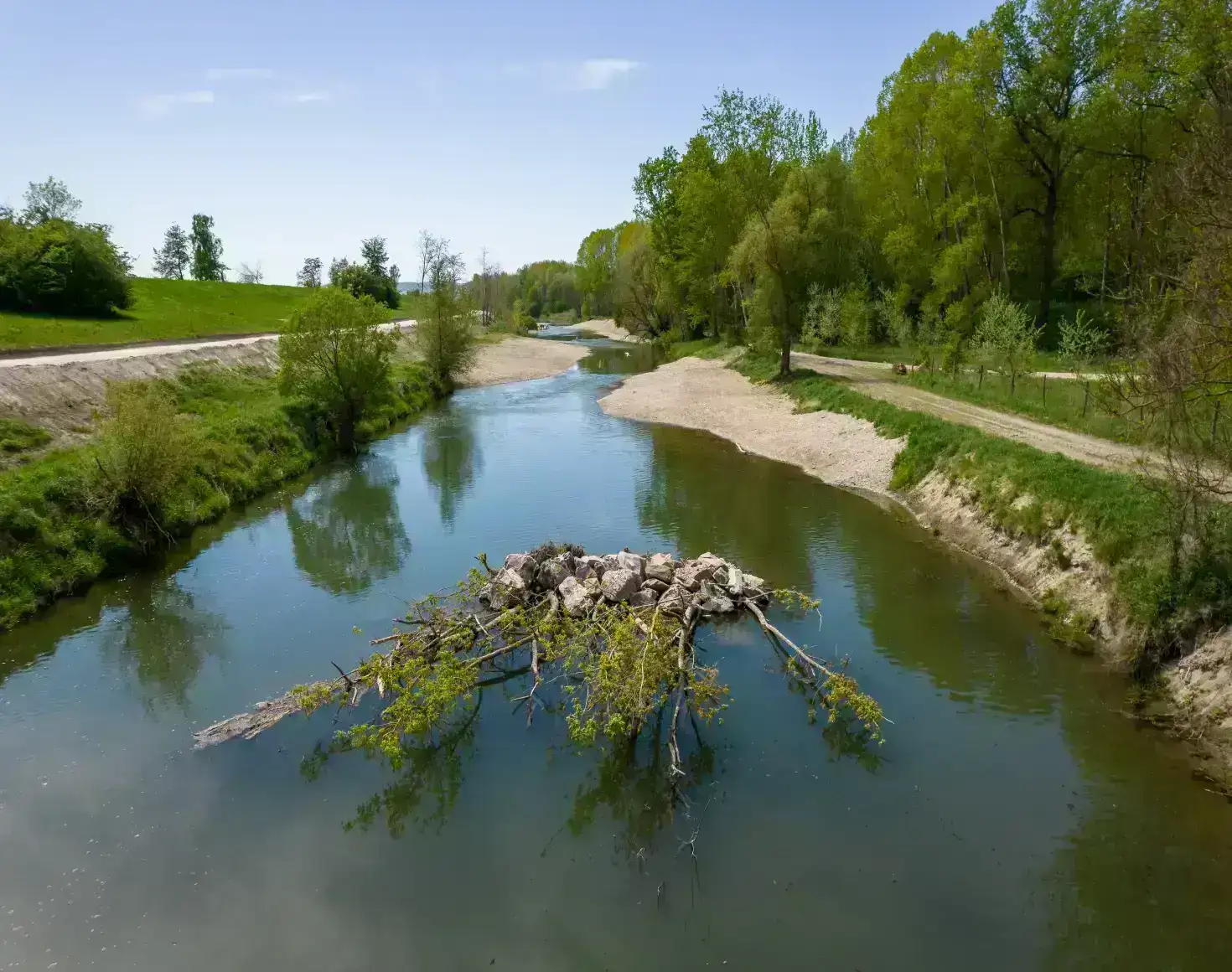 Das Bild zeigt eine renaturierte Stelle bei der Fischwanderhilfe in Altenwörth. Im blaugrünen Fluss befindet sich an dieser Stelle eine Ansammlung von Steinen mit Pflanzen, wie man sie auch in der Natur findet. Links und rechts an den Ufern sieht man aufgeschütteten Kies sowie Grünflächen mit Sträuchern und Bäumen. Im oberen Bildbereich strahlt der blaue Himmel.