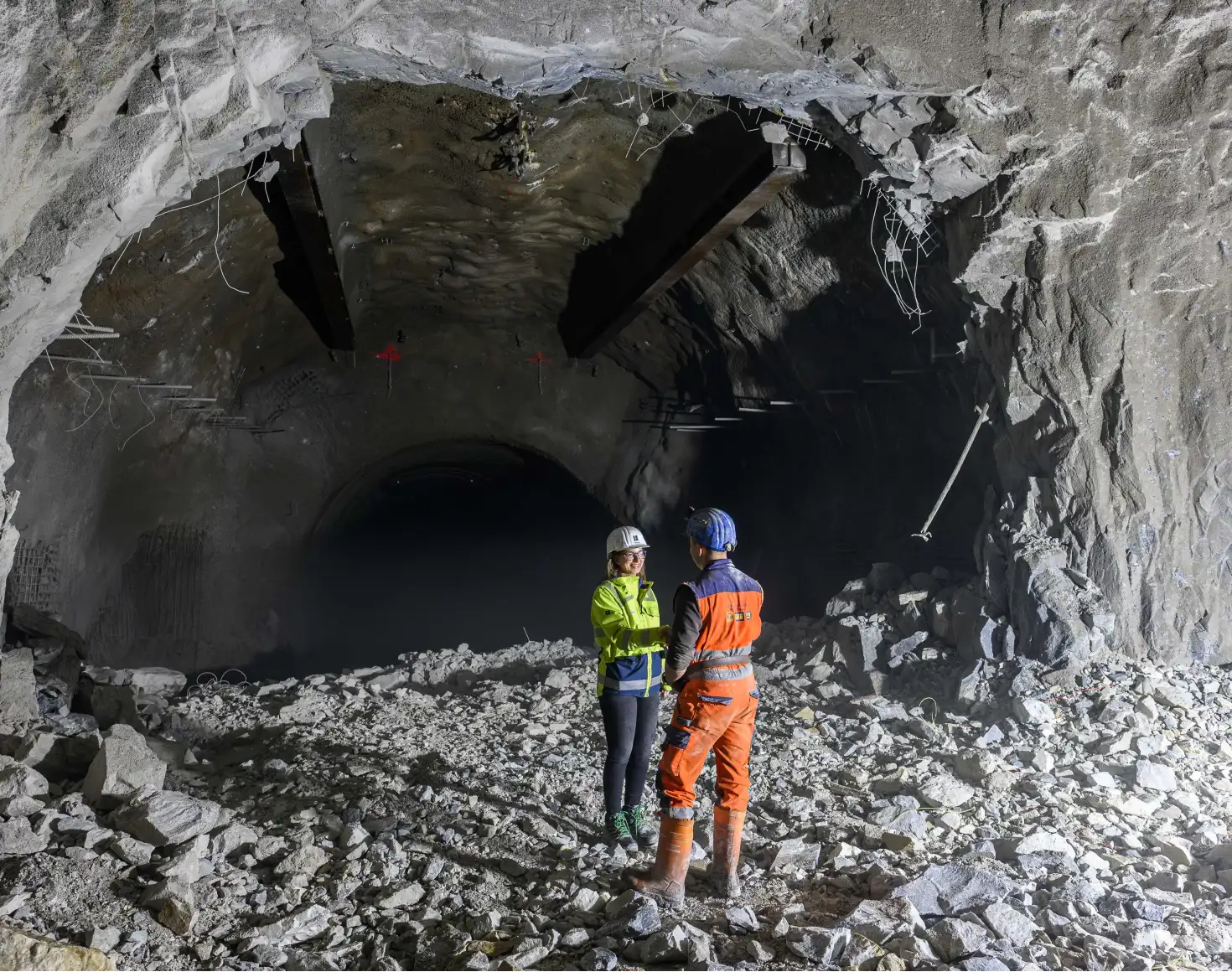 VERBUND-Projektleiterin Maria Leo in einer gelben Sicherheitsjacke und Helm gratuliert einem Mineur in oranger Arbeitskleidung und blauem Helm im Inneren eines Tunnelbaustelle. Im Hintergrund sieht man die offene Tunnelwand und Steinschutt auf dem Boden.