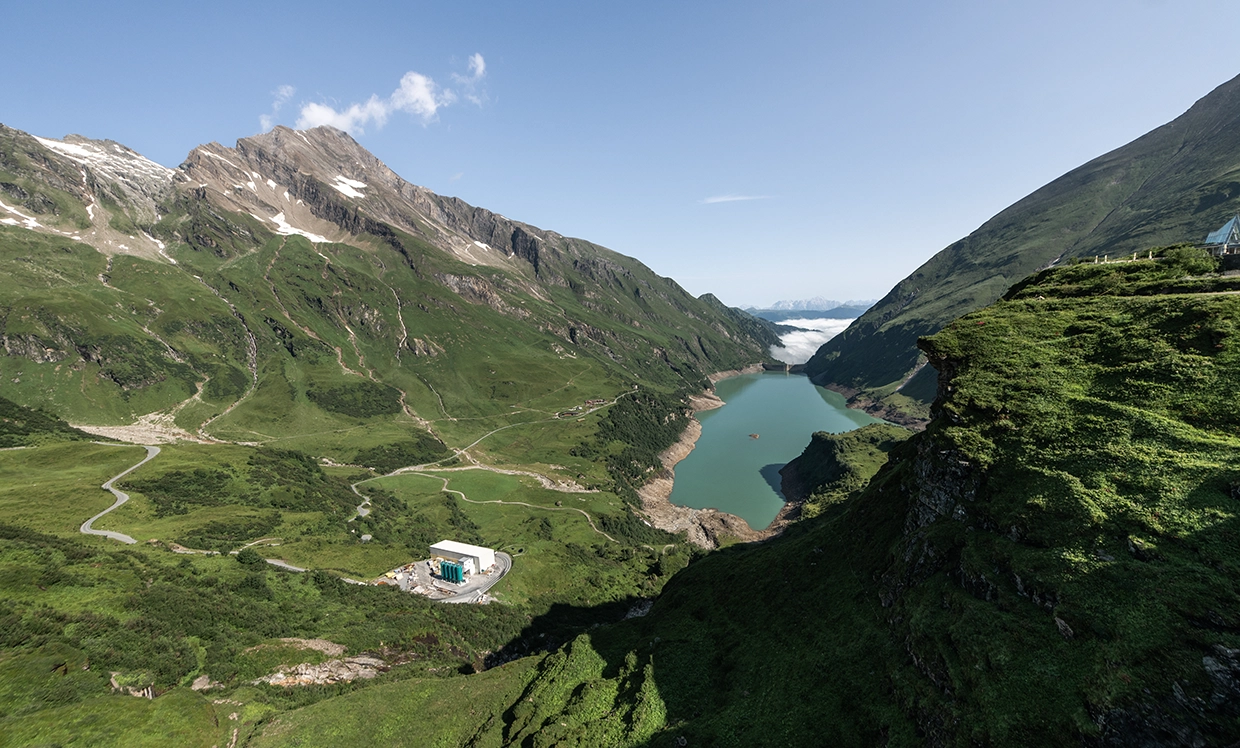 Ein Blick von oben auf den Stausee Mooserboden in Kaprun. Der Himmel ist blau und die Sonne scheint. Der Stausee ist strahlend blau.