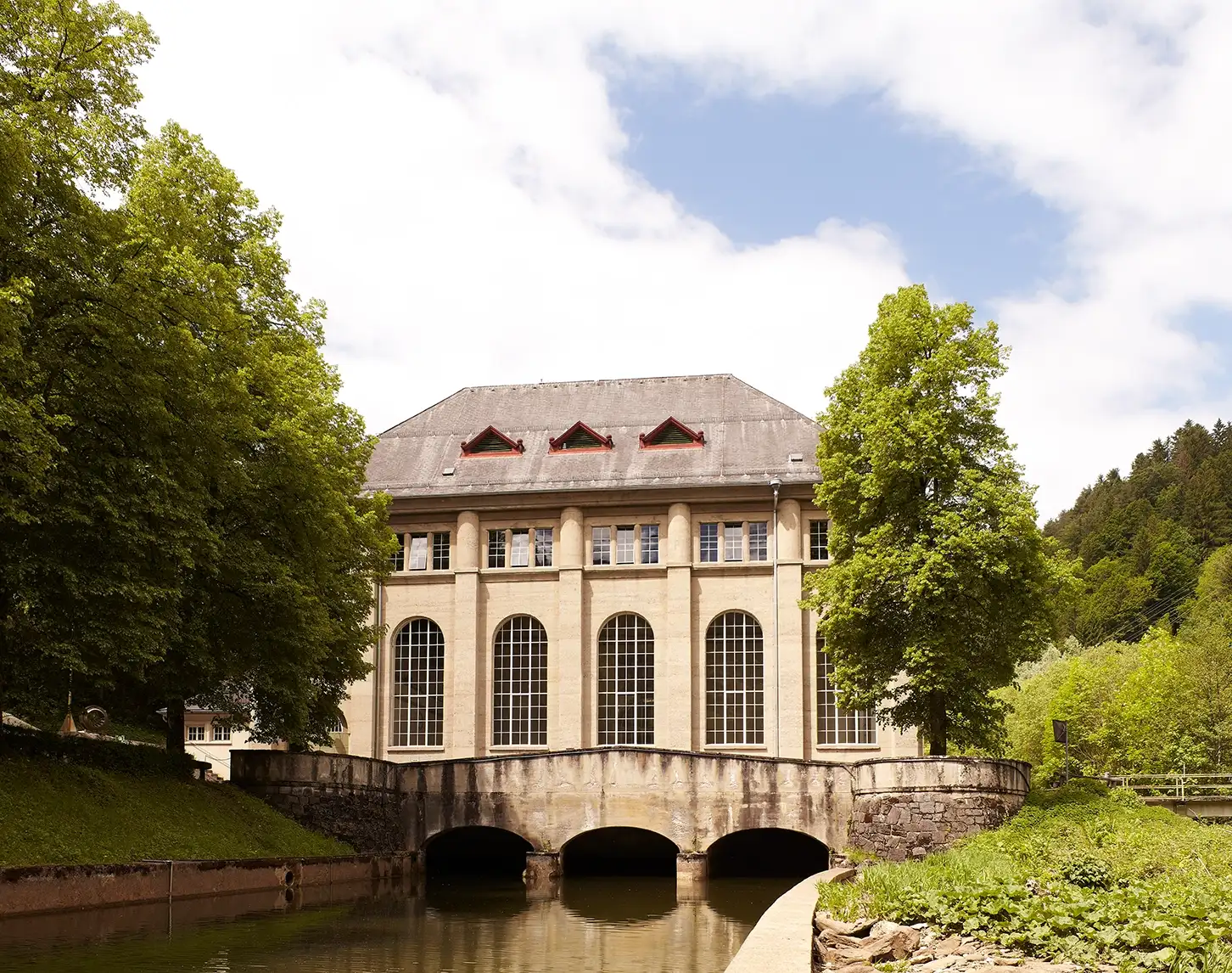 Wir blicken auf das Wasserkraftwerk in Arnstein. Das Gebäude besteht aus hellbraunen Steinen. Davor sehen wir eine kleine Brücke. Rechts und links sehen wir Bäume, Wiese und Sträucher.