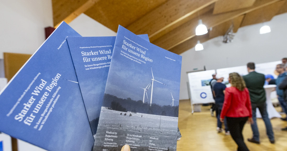 Mehrere Exemplare einer Projektzeitung mit dem Titel „Starker Wind für unsere Region“ werden gehalten. Die Zeitung beschäftigt sich mit dem Windkraftprojekt Burgschleinitz-Kühnring. Im Hintergrund sind Personen in einem Veranstaltungsraum zu sehen.