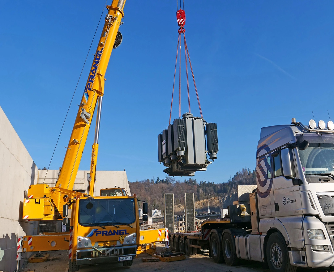 Ein großer Transformator wird an einem sonnigen Tag mit einem gelben Mobilkran auf einem Baustellengelände installiert. Der Transformator schwebt in der Luft, gesichert mit roten Hebebändern, während er behutsam über einen Lastkraftwagen, auf dessen Anhänger er zuvor transportiert wurde, manövriert wird. Im Hintergrund ist ein klarer blauer Himmel über einem hügeligen, bewaldeten Horizont sichtbar. Baugeräte und Sicherheitszäune sind um den Installationsbereich herum angeordnet, was auf eine gut organisierte und aktive Baustelle hindeutet.