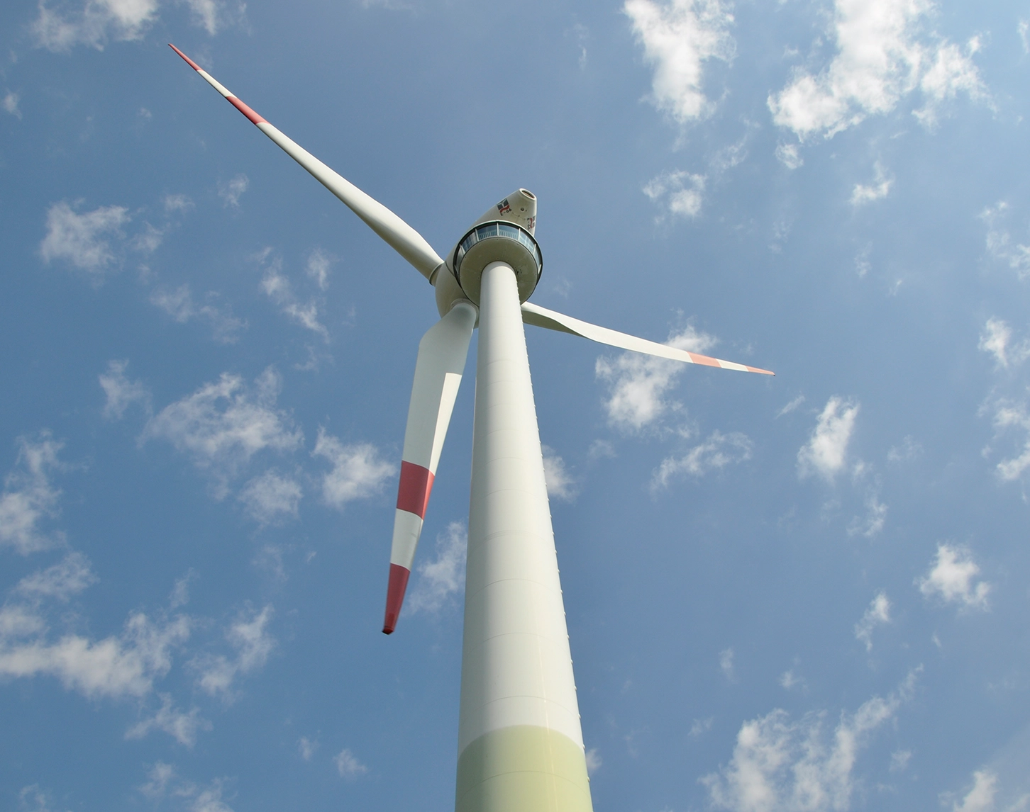 Wir blicken von unten aufs Schauwindrand in Bruck an der Leitha. Der Himmel ist blau und man sieht kleinere Wölkchen. Das Windrad ist größtenteils weiß. Die Spitzen der Windräder sin rot. Unterhalb der Turbine sieht man die Besucherplatzform.