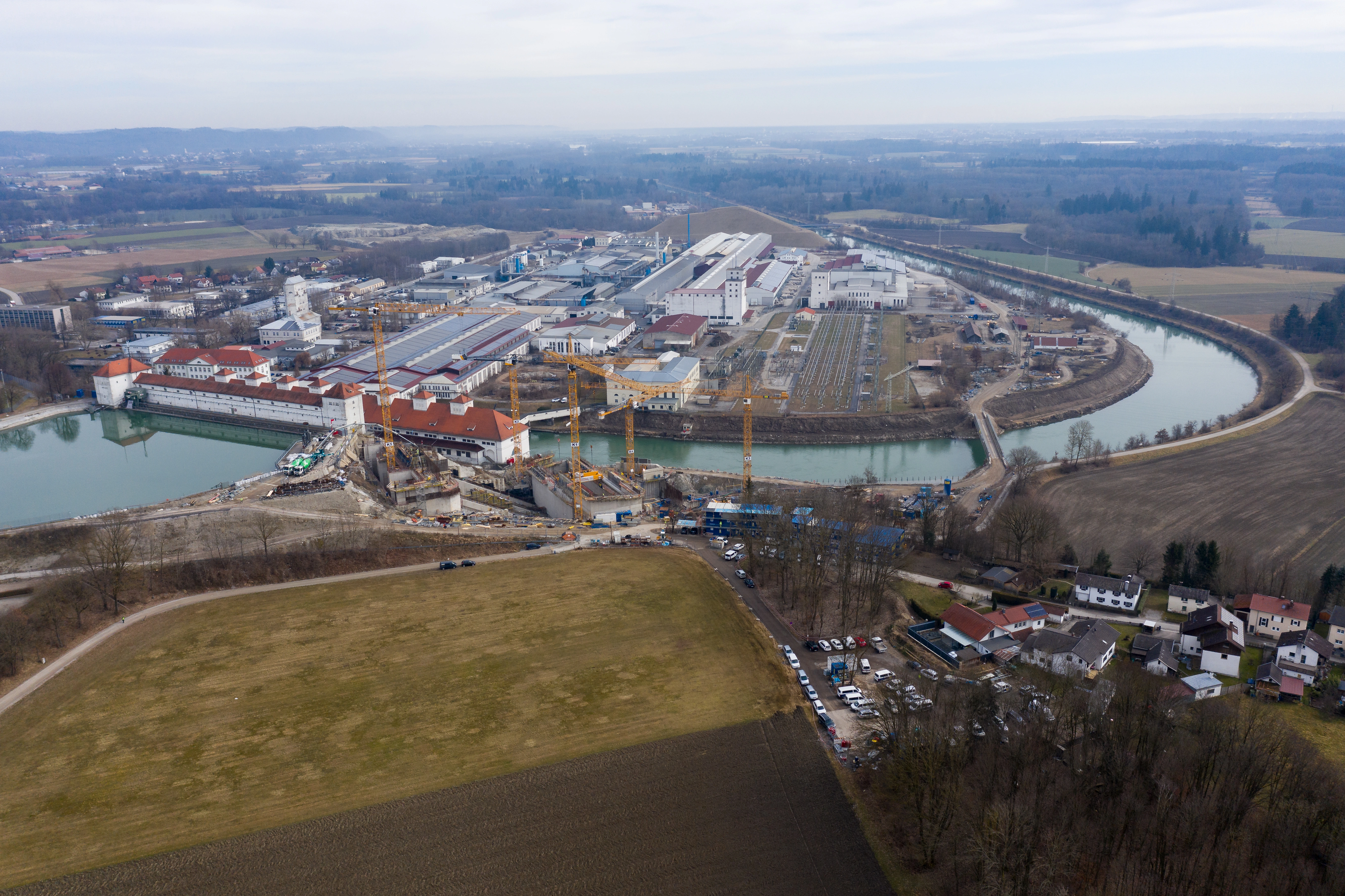 Luftaufnahme der Baustelle zur Erneuerung des VERBUND Kraftwerks Jettenbach-Töging. Mehrere Kräne und Bauarbeiten sind entlang eines Flusslaufs sichtbar, umgeben von industriellen Gebäuden und landwirtschaftlichen Flächen.