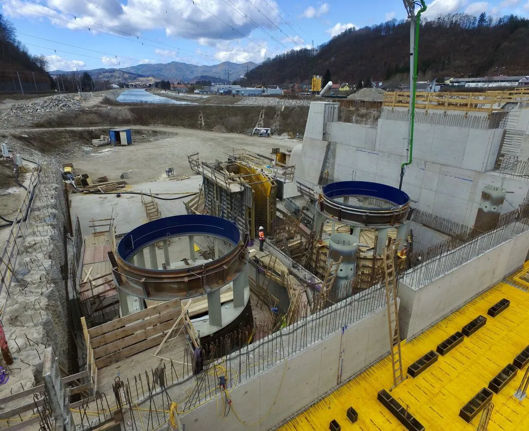 Ein Bild der Baustelle zeigt die zwei großen runde Turbinenschächte, umrandet von blauen Schutzabdeckungen, bevor die Turbinen eingebaut wurden. Die Schächte ragen aus einem Betonfundament heraus, das von zahlreichen Stützstreben und Baugerüsten umgeben ist. Im Vordergrund ist eine gelbe Schalung zu sehen, die als Basis für weitere Betonarbeiten dient. Ein Arbeiter in Schutzbekleidung steht zwischen den Schächten und betont die Größendimension der Struktur. Im Hintergrund erstreckt sich eine weitläufige Baustelle unter einem bewölkten Himmel. Berge und eine Flusslandschaft sind in der Ferne sichtbar.