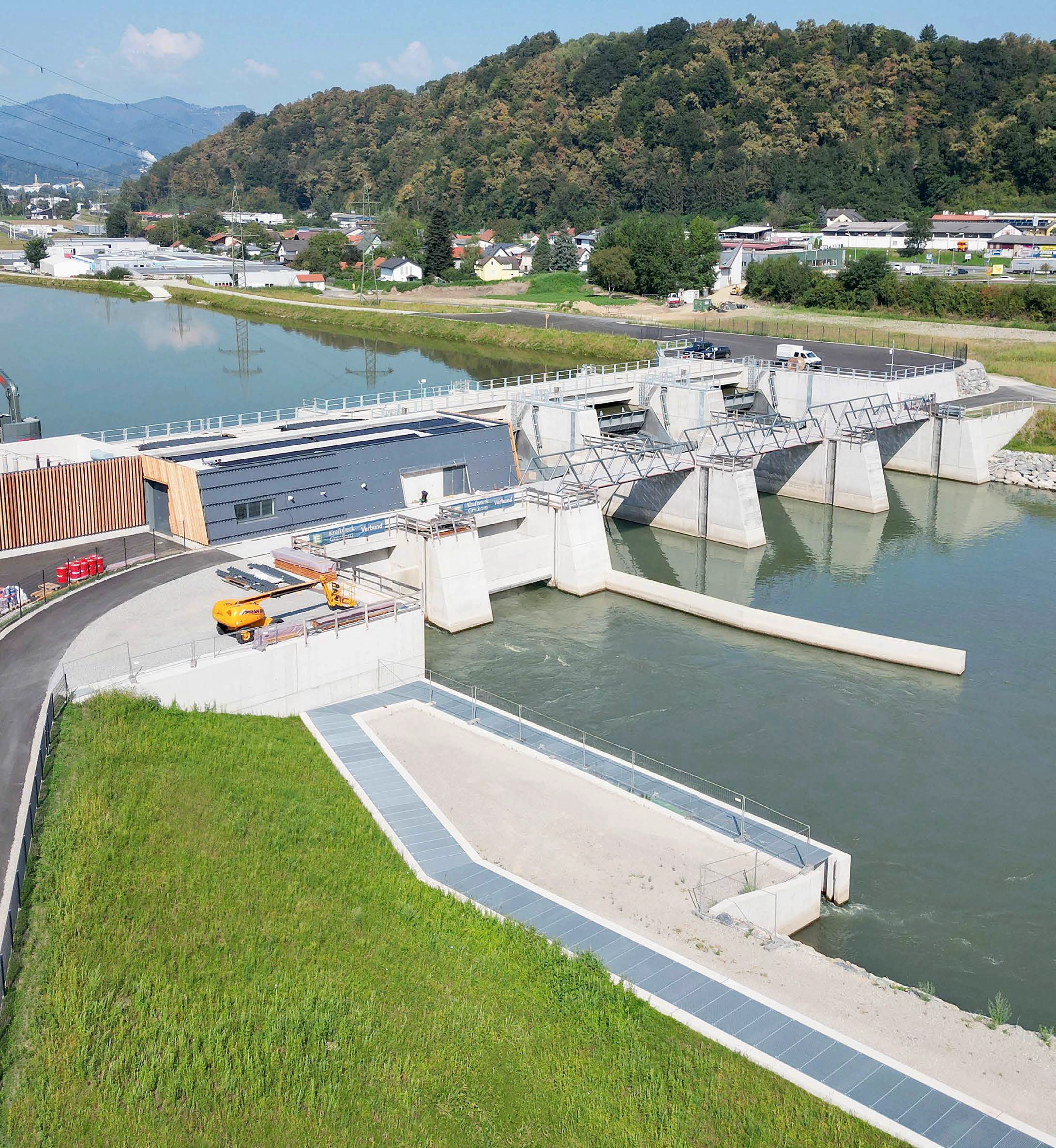 Luftaufnahme des neu errichteten Murkraftwerks Kraftwerk Gratkorn. Man sieht den Wasserdurchlass, das Betriebsgebäude und das Wehr mit geöffneten Schleusentoren in einer grünen Landschaft mit einem Fluss.
